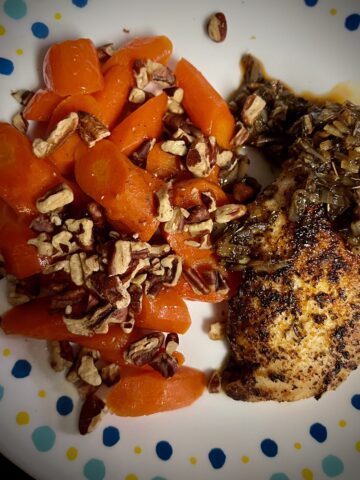 Pecans covering carrots on the right side of the plate have a nice shine to them chicken on the right side covered in seasoning and has nice sauce and shallots for a nice chicken Demi glaze with glazed carrots