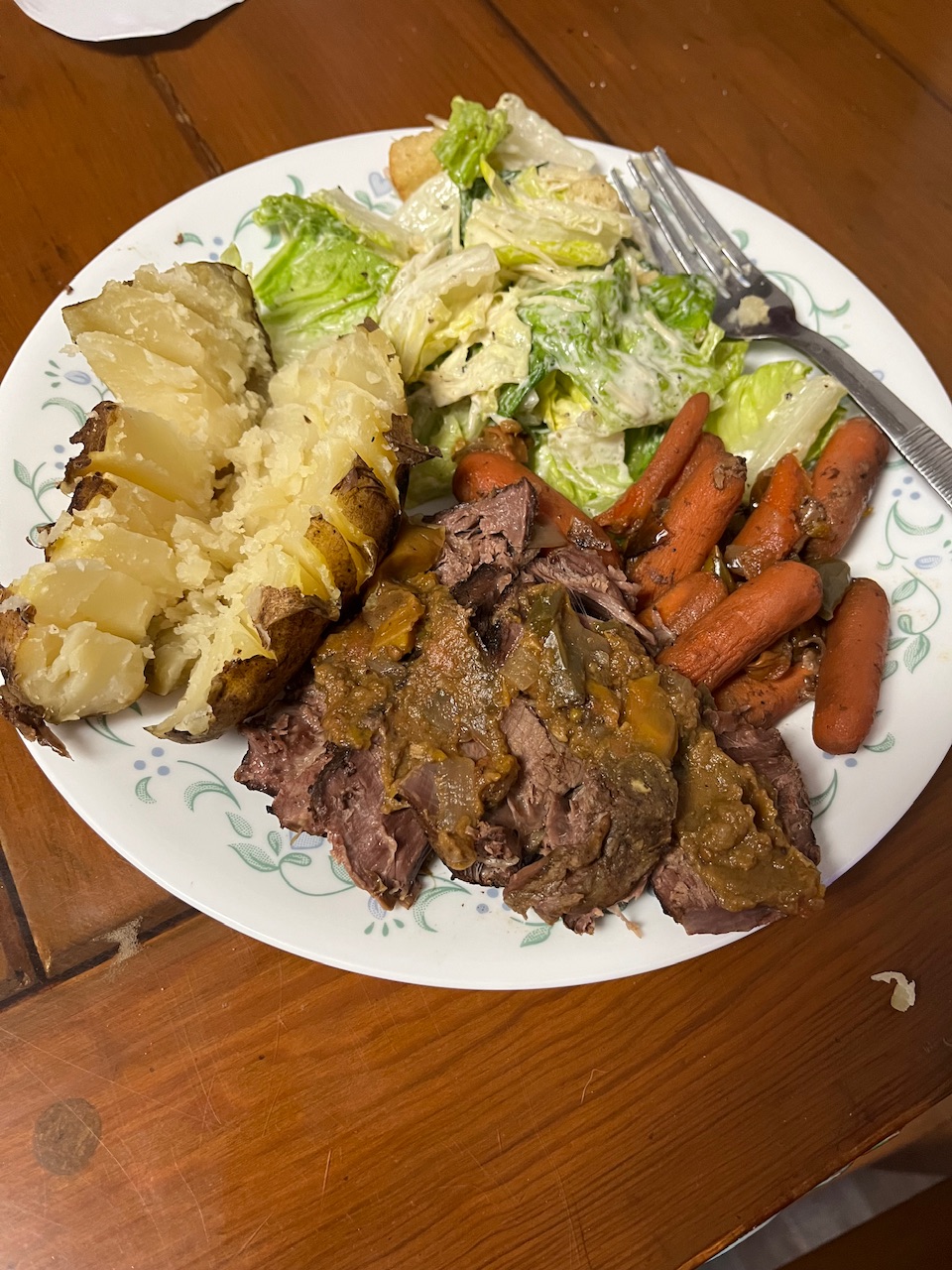 Some slice of roast have been placed on a plate that’s sitting on a round table. Potato has been sliced in half and is to the right above the roast along with some salad that is upper right roast.